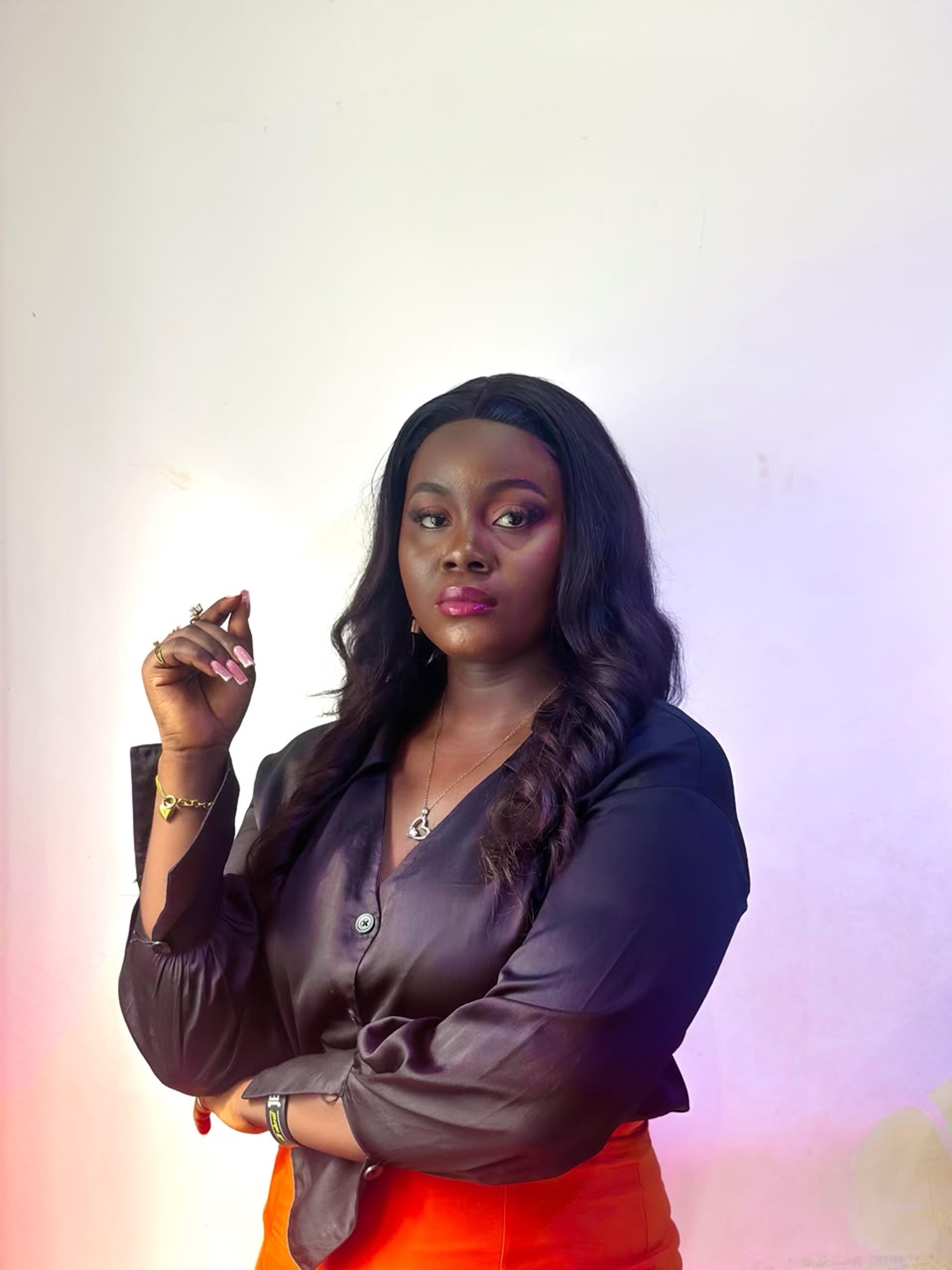 Omena Otovo — portrait, one arm folded and the other hand raised, in a dark satin blouse and orange trousers against a pale wall washed in warm pink light.
