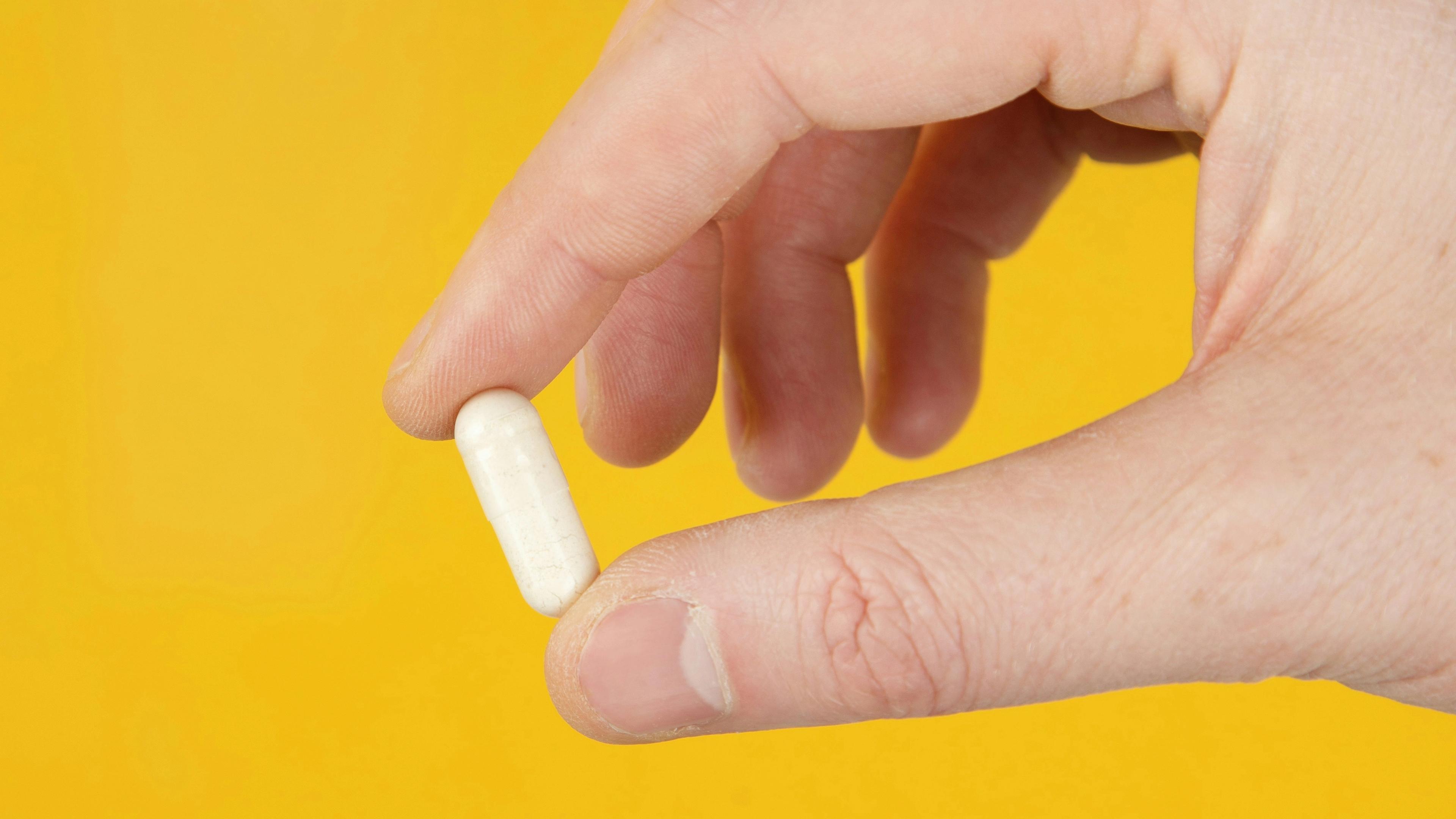 A hand holding a white pill against a yellow background