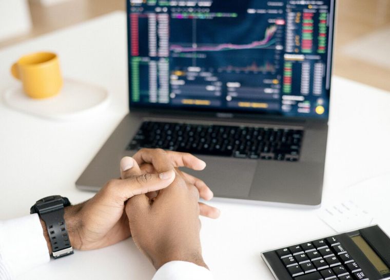 A person sits at a desk with hands clasped, facing a laptop displaying colorful stock market charts and data, alongside a calculator—illustrating the analysis and strategic planning involved in ESG investing to potentially enhance returns and leverage tax-saving opportunities.