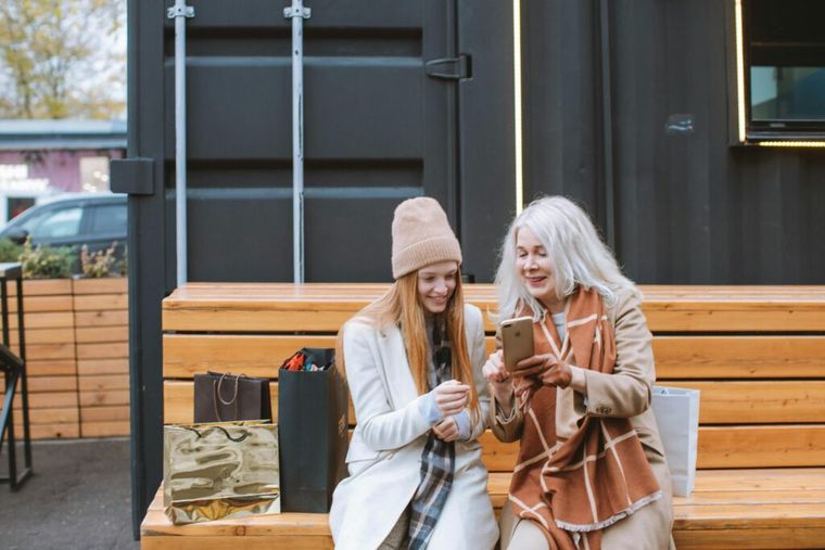 Retired woman sitting on a bench outdoors with her daughter while they both look at the older woman’s iPhone.