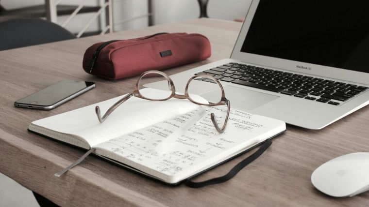 Workspace setup on a wooden desk featuring an open notebook with handwritten notes, a pair of round eyeglasses resting on top, a white pen, a closed red pencil case, a MacBook Air laptop, a smartphone, and an Apple Magic Mouse.