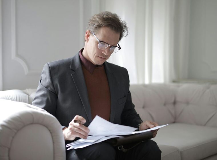 A classy executive male reading financial documents while seated on a couch, symbolizing the importance of retirement planning.