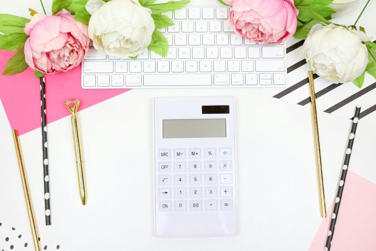 Bright and colorful flat lay of a feminine workspace featuring a white calculator in the center, a white keyboard partially visible at the top, pink and white peonies, gold pens, black-and-white polka-dotted straws, and pink and patterned paper backgrounds. The scene has a cheerful, creative aesthetic.