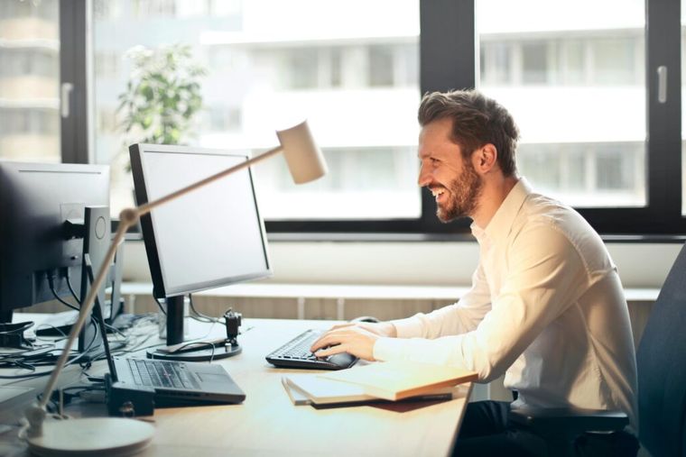 Smiling man in a white dress shirt sitting in an office chair, facing a computer.