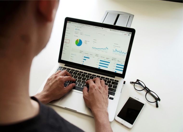 Person analyzing website analytics on a laptop, with various graphs, charts, and data visible on the screen. The individual is typing with both hands, and nearby on the white desk are a smartphone and a pair of black-rimmed glasses. The setting suggests a focus on digital marketing or data analysis.