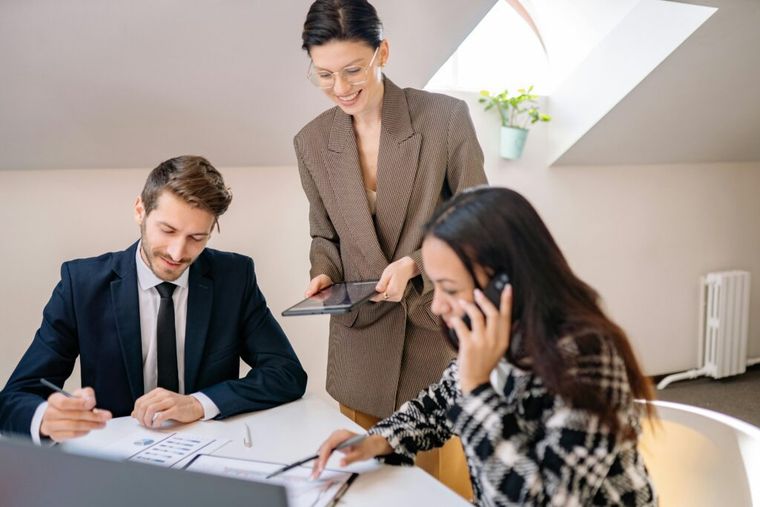 Three business professionals collaborating around a table—one seated reviewing documents, one standing presenting with a tablet, and one speaking on the phone—in a modern office meeting.