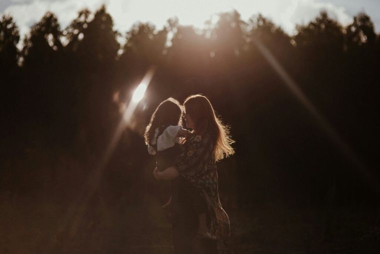 A woman holding her toddler, outside with the sun shining down.