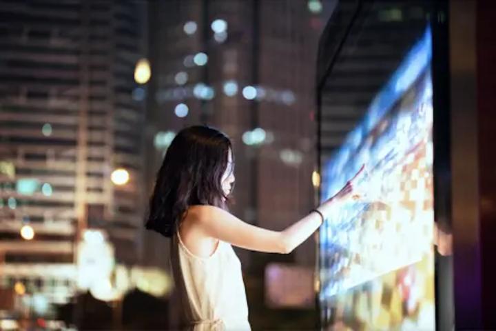 Person using a large interactive digital display inside a glass-walled urban space at night