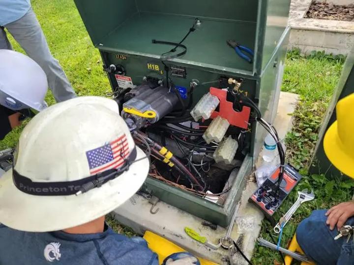Utility workers inspecting an open electrical control cabinet with DTM, wiring, tools, and safety helmets