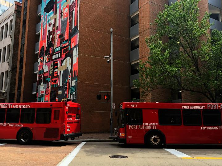 Two red transit buses stopped along a downtown pittisburgh street beside a mural-covered building and below a street light pole with UbiHub