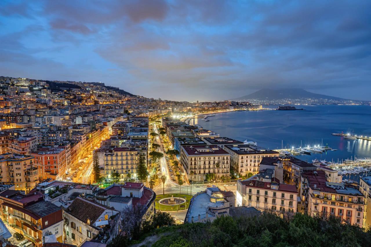 Elevated view of a coastal city at dusk with illuminated streets, buildings, and a marina along the shoreline