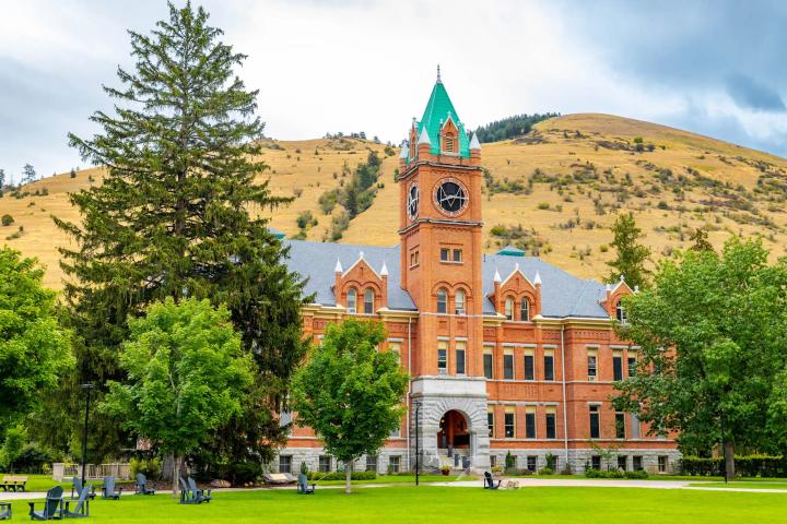 Brick building with central clock tower surrounded by trees and grassy lawn in a mountainous campus setting