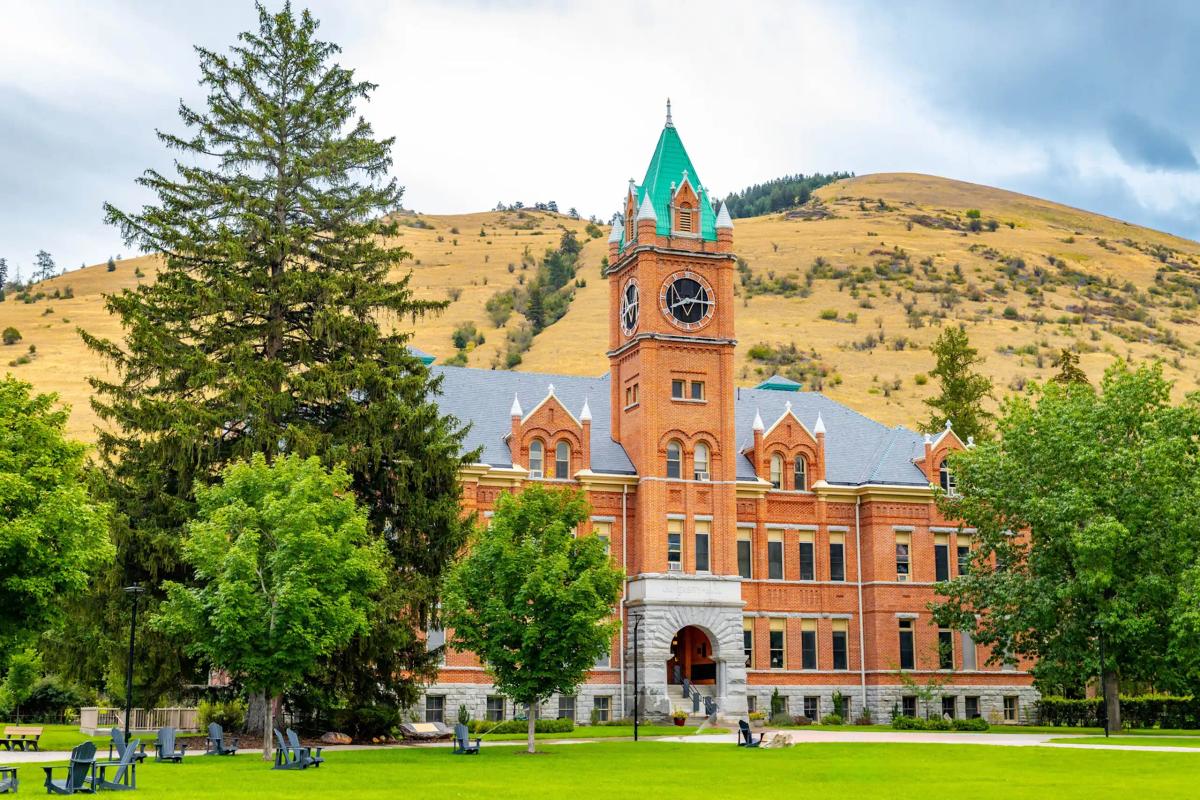 Brick building with central clock tower surrounded by trees and grassy lawn in a mountainous campus setting