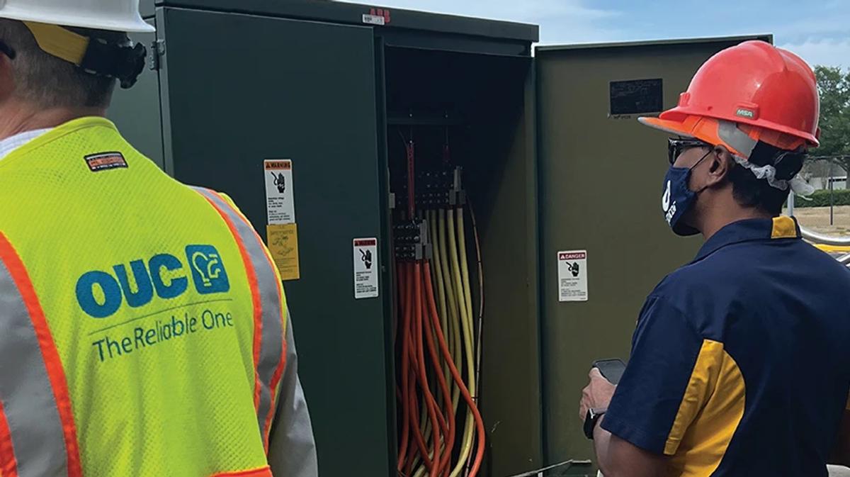 Two workers wearing safety gear inspect open electrical cabinet with multiple power cables outdoors
