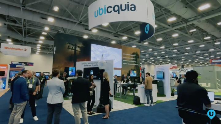 Ubicquia exhibition hall booth with overhead company signage, display screens, and attendees talking on the show floor