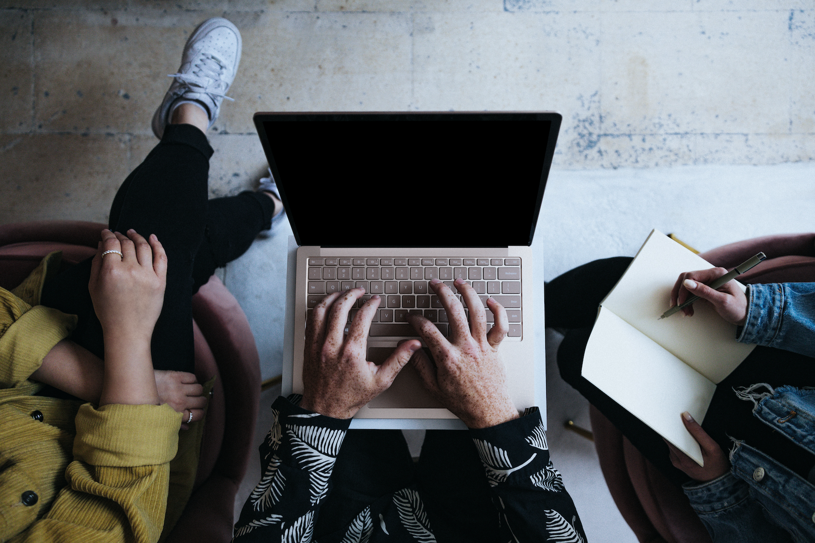 An aerial shot of three people sitting together, one taking notes and another typing on a laptop.