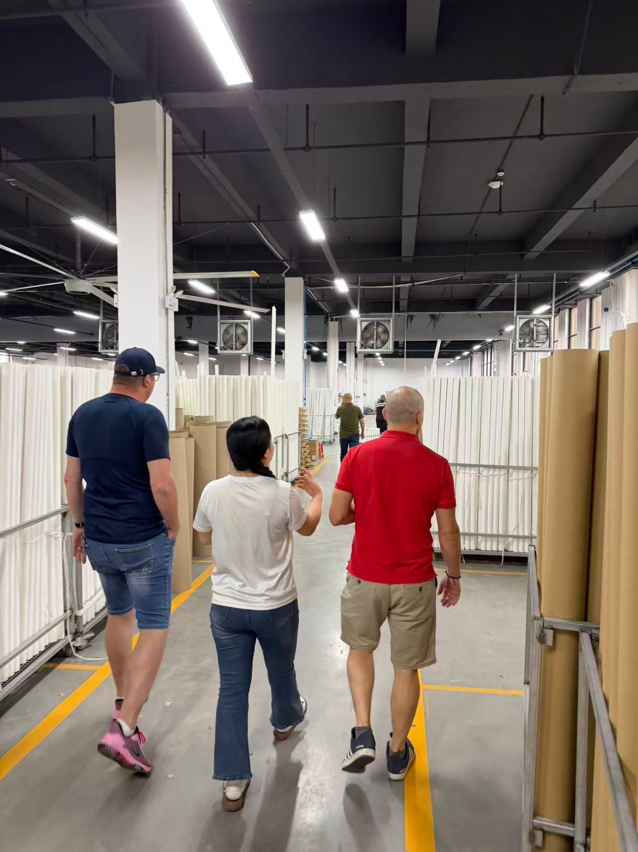 Foreign buyers follow factory manager to inspect rolls of white upholstery lining non-woven fabric in furniture factory warehouse, these textile base cloths are inner padding materials for modular sofas, not plastic rolls