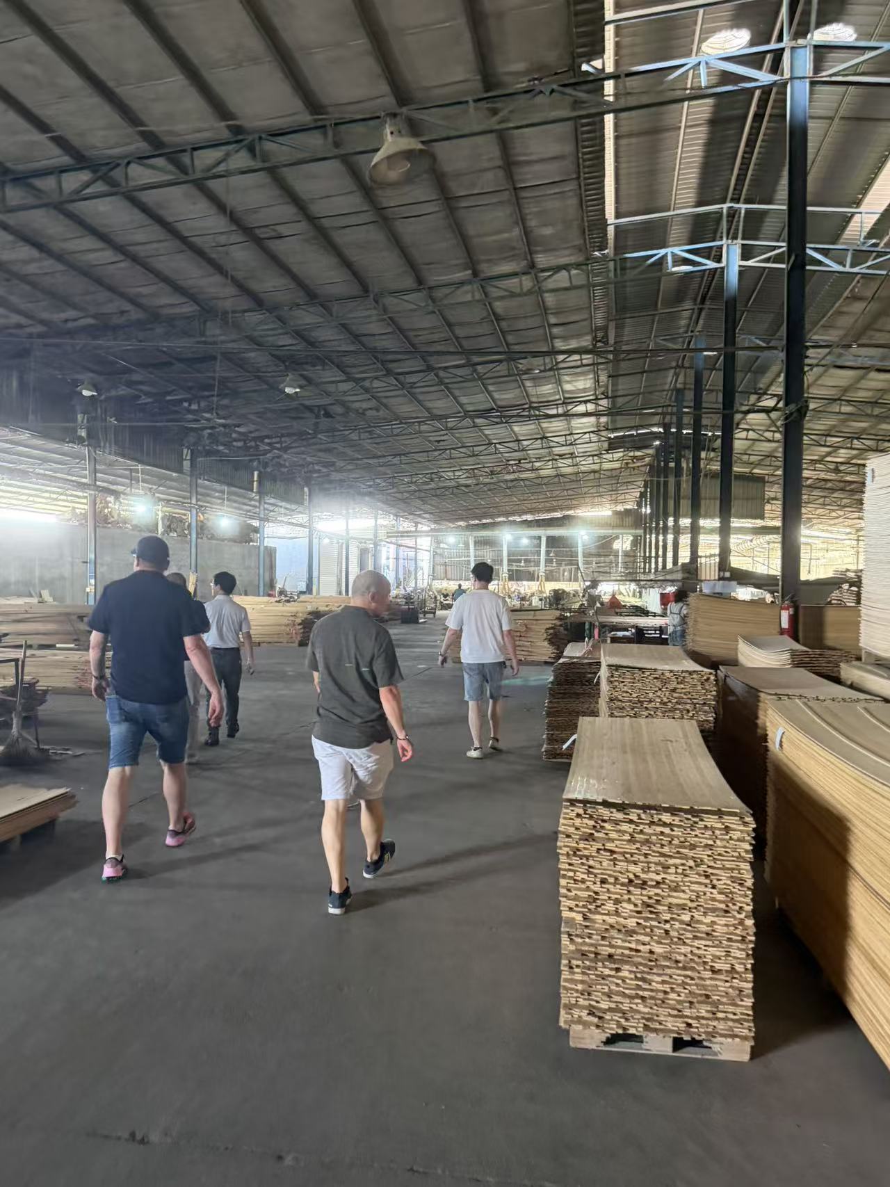 Overseas purchasing team walk through timber raw material storage workshop during China furniture factory inspection, buyers check stacked wood veneer and plywood raw materials for upholstered sofa production