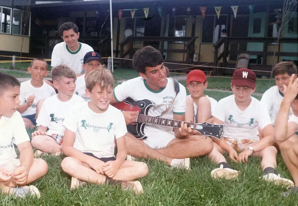 vintage photo of a group of young men sitting on top of a lush green field surrounding a man with a guitar