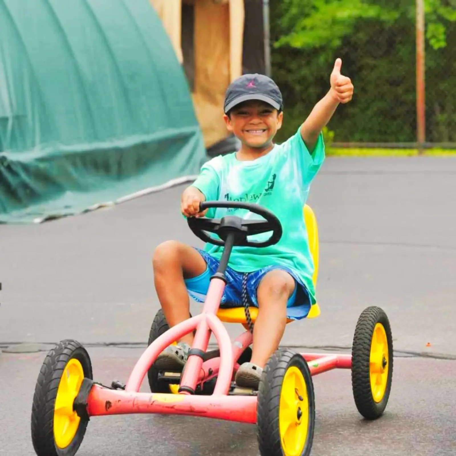 A smiling boy in a Maplewood shirt giving a thumbs up while riding a pedal kart