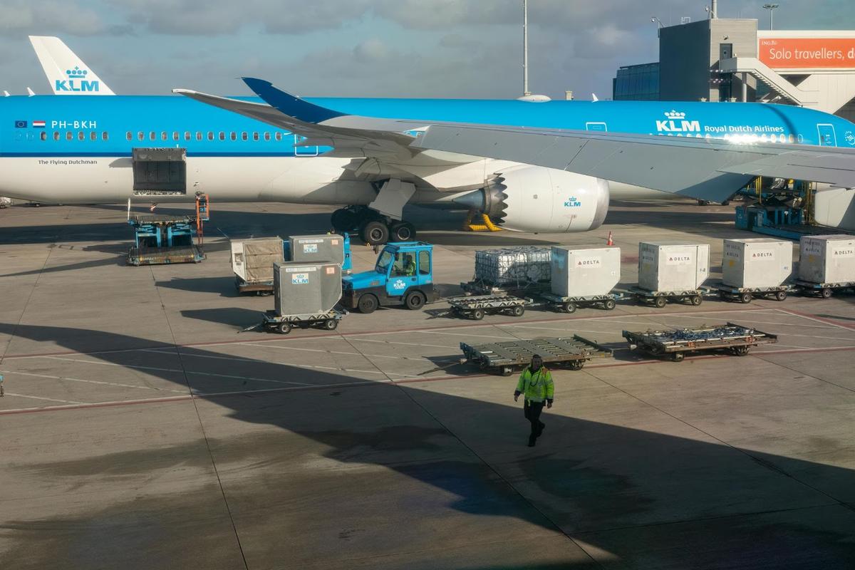 空港ゲートに着陸したKLM機が、貨物の積み込みと地上ハンドリング作業を行っています。航空会社のロゴが入ったコンテナが積み込み準備のために配置され、地上クルーが積極的に物流を管理しています。