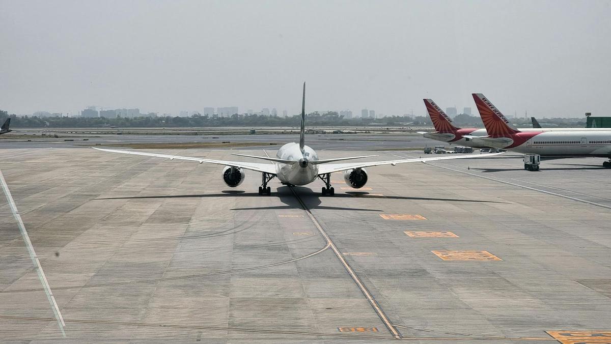 A rear view of a modern commercial jet taxiing at an international airport, flanked by two international passenger aircraft. A hazy skyline and wide tarmac reflect the growing scale and complexity of contemporary global aviation operations.