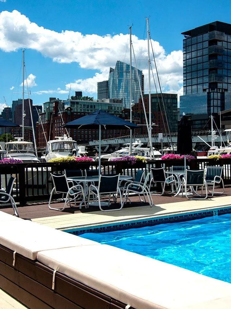 A pool area with a deck, tables, chairs, and umbrellas, overlooking a city skyline.