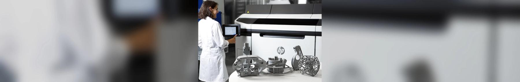 A technician in a lab coat operates the touchscreen of an HP Multi Jet Fusion 3D printer, with several finished additive-manufactured parts displayed on the table in front of the machine.