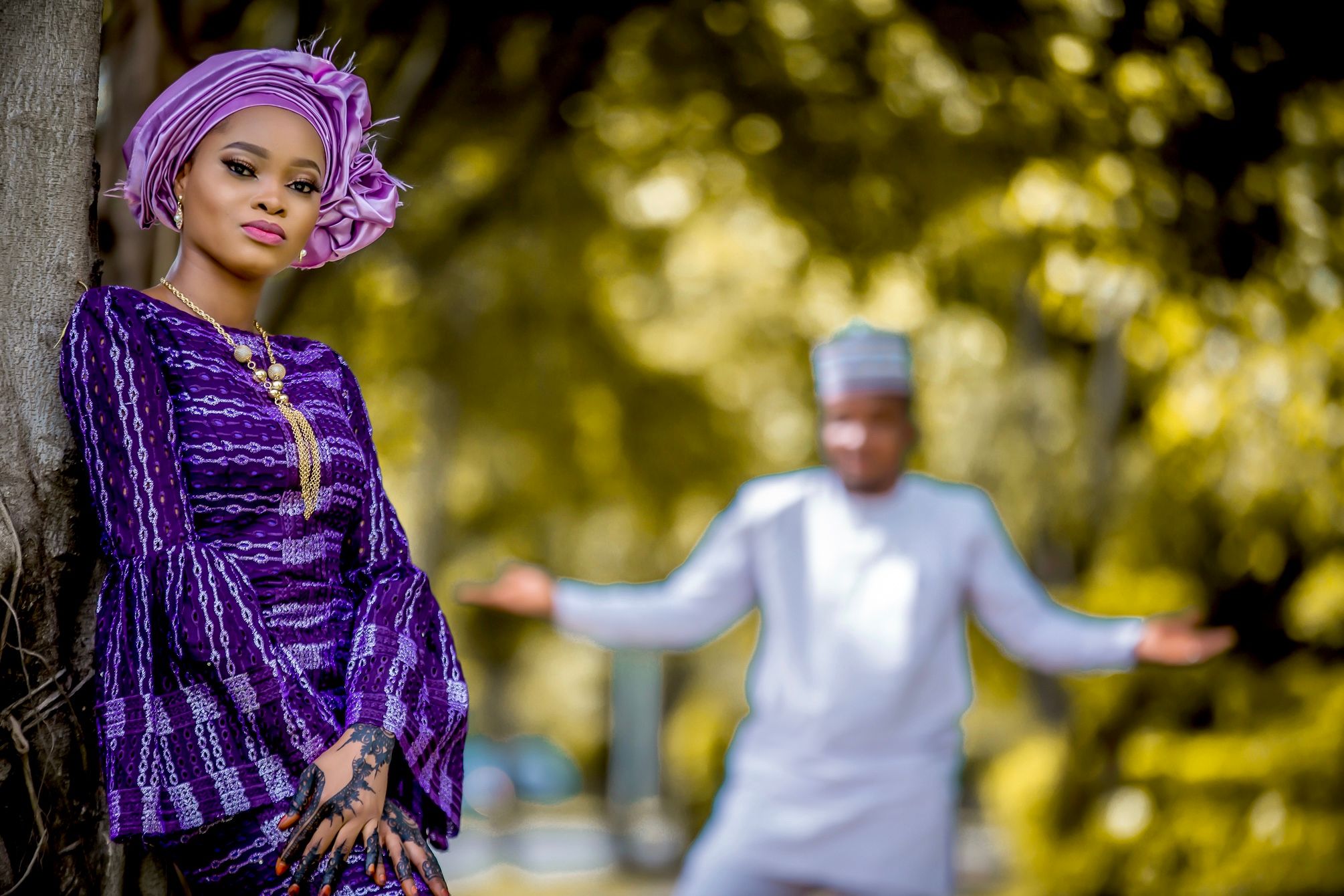 Bride in a purple gele and lace leaning against a tree, the groom out of focus behind her