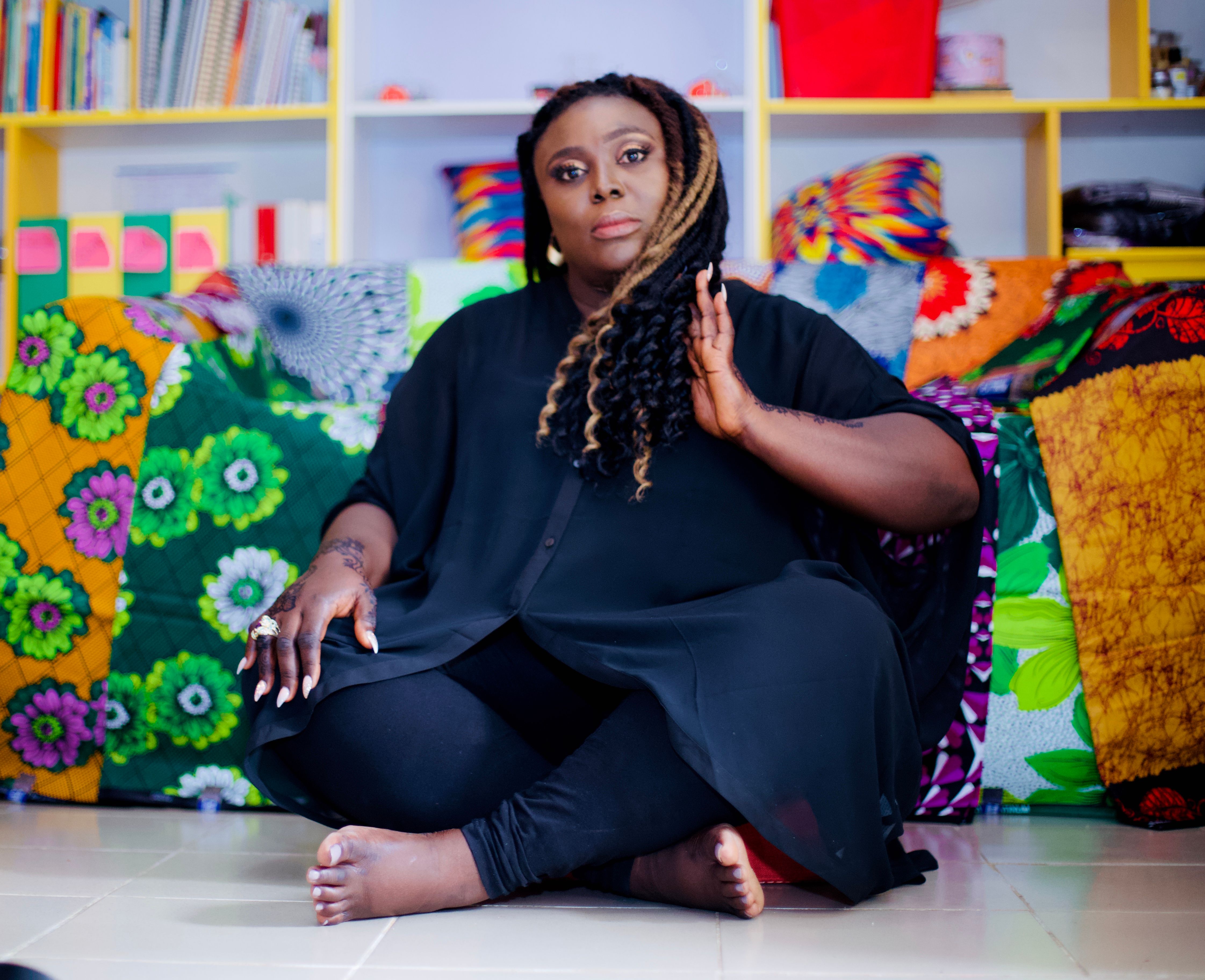 Woman in black seated among brightly patterned cushions, hand raised to her braids