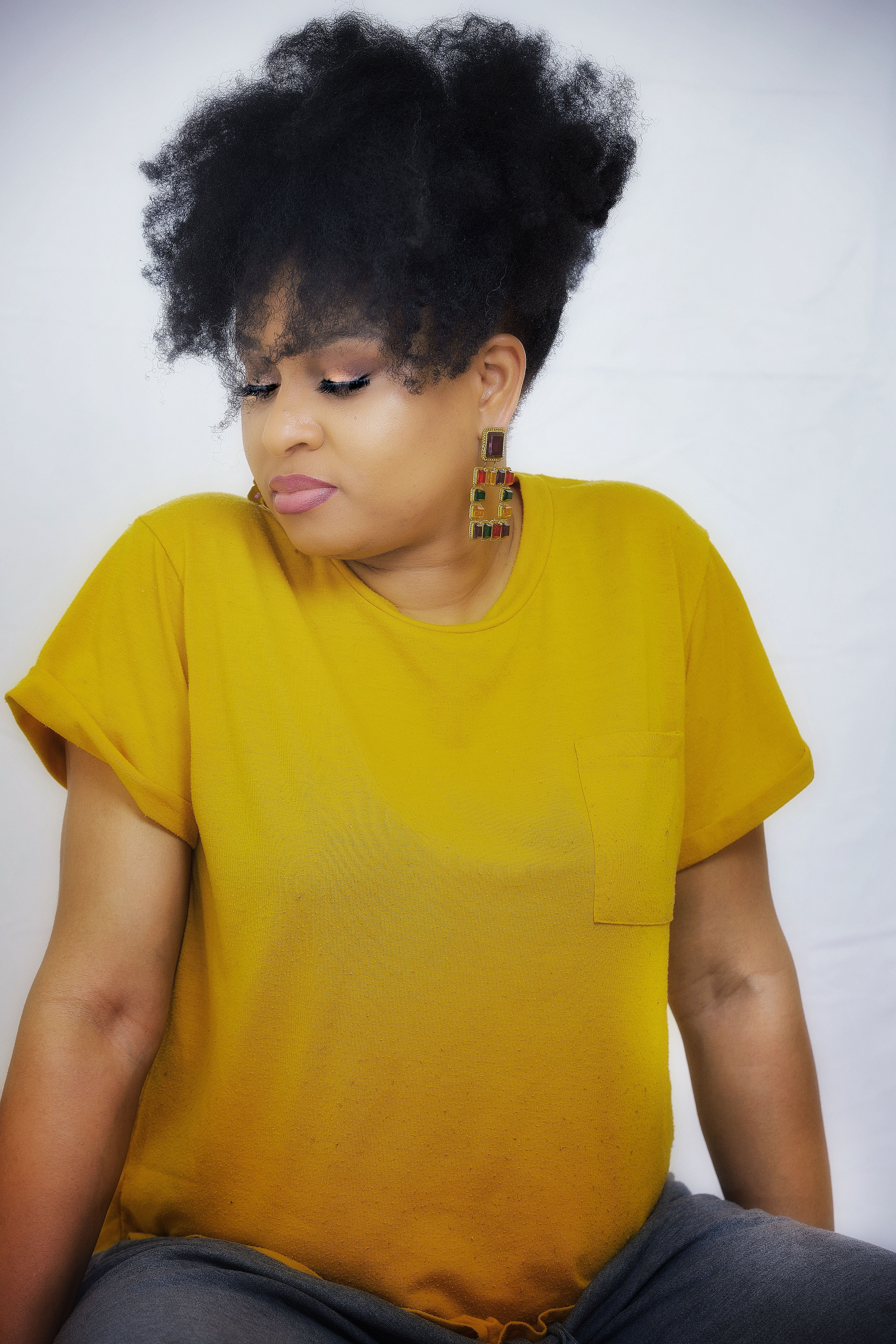 Woman in a mustard t-shirt with an afro updo and beaded earrings, looking down