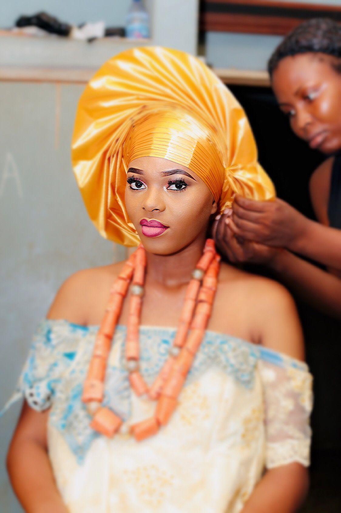 Bride in a gold gele and coral beads as her headwrap is tied