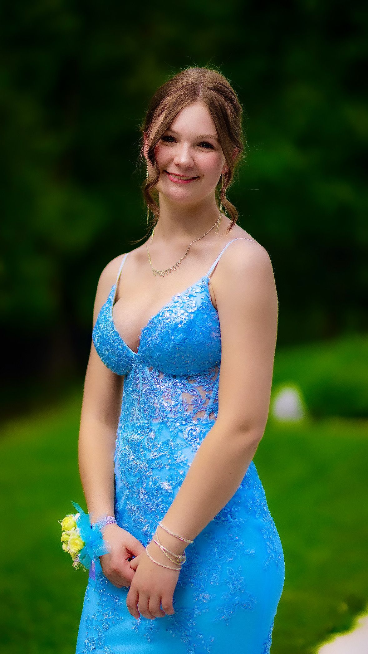 Graduate in a blue lace gown with a corsage, smiling in a garden