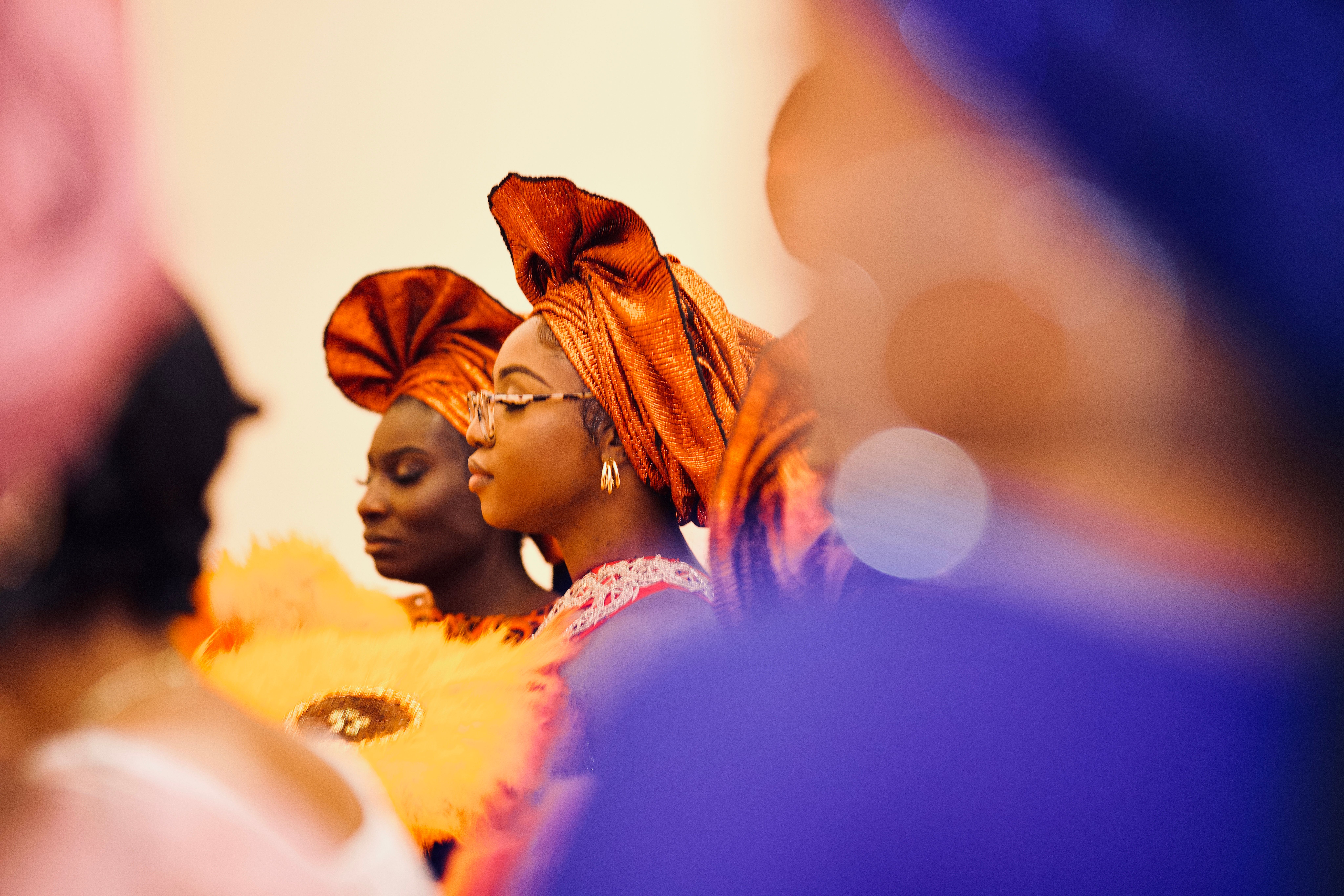 Two guests in copper gele headwraps in profile, shot through a blurred foreground