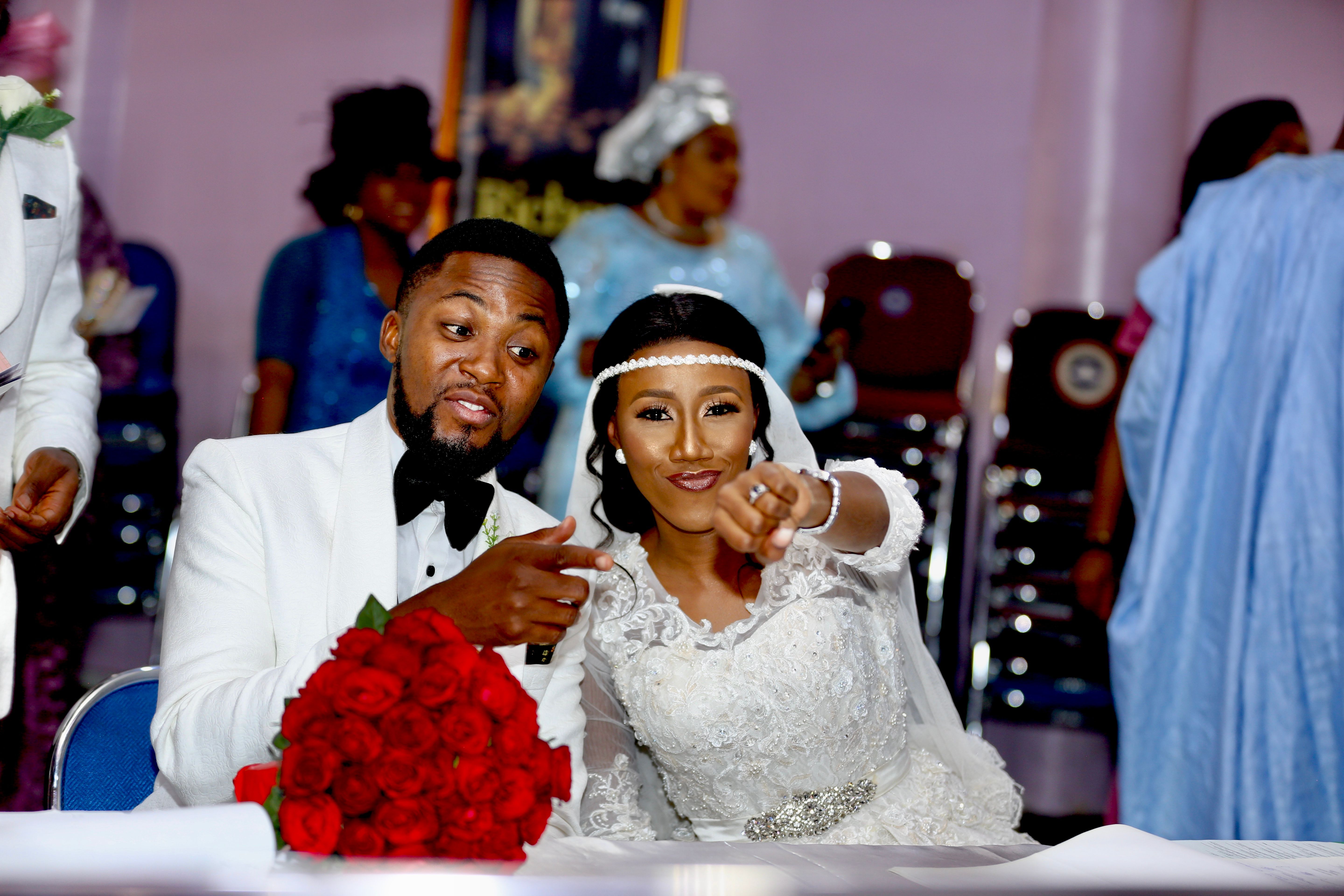 Bride and groom at the signing table, both pointing at the camera behind a bouquet of red roses