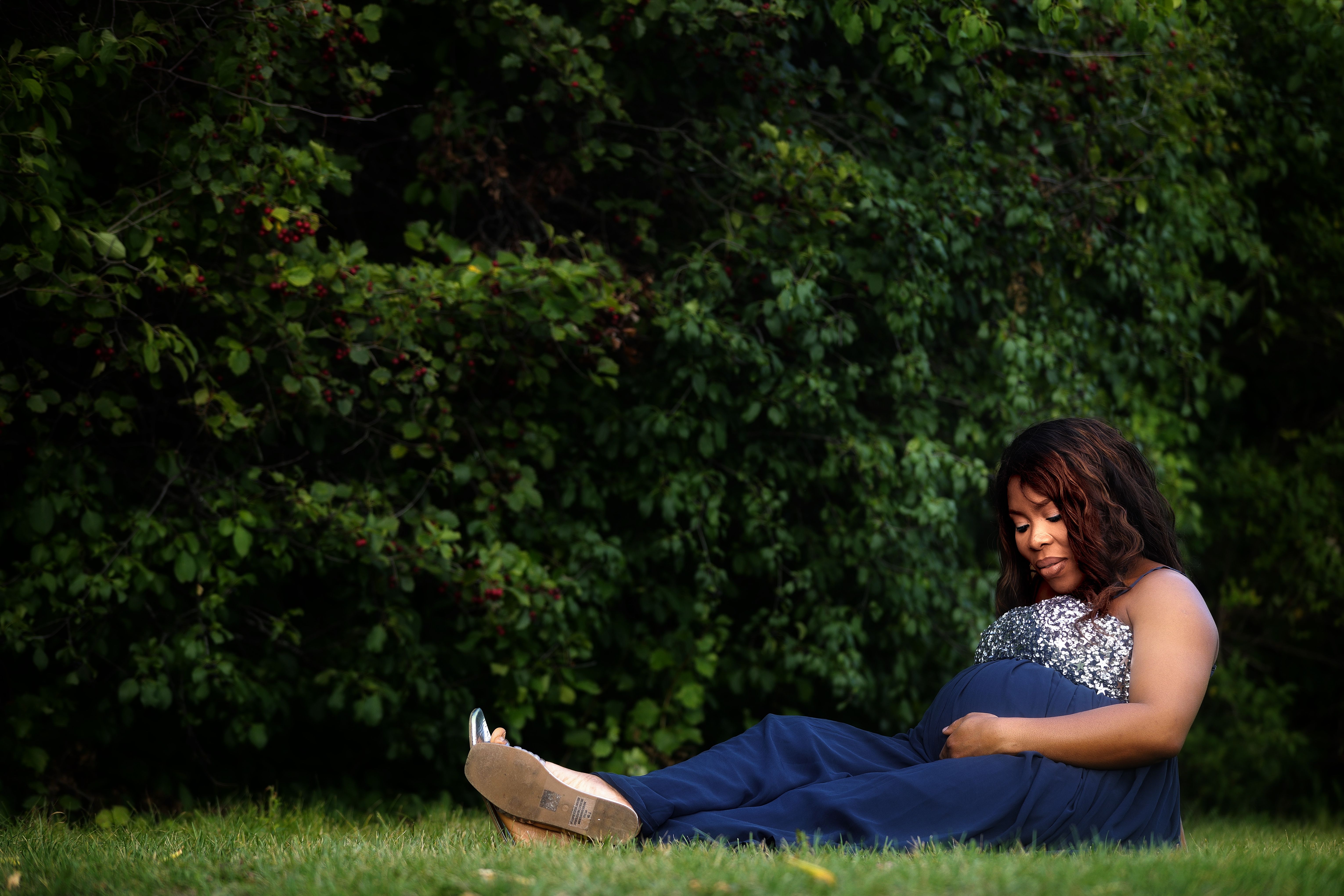 Maternity portrait in a navy gown, seated on the grass beneath a berry tree