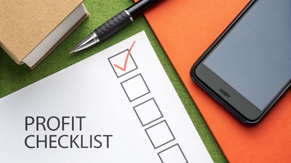 Overhead view of a 'PROFIT CHECKLIST' document with a red checkmark, next to a pen, book, and smartphone.