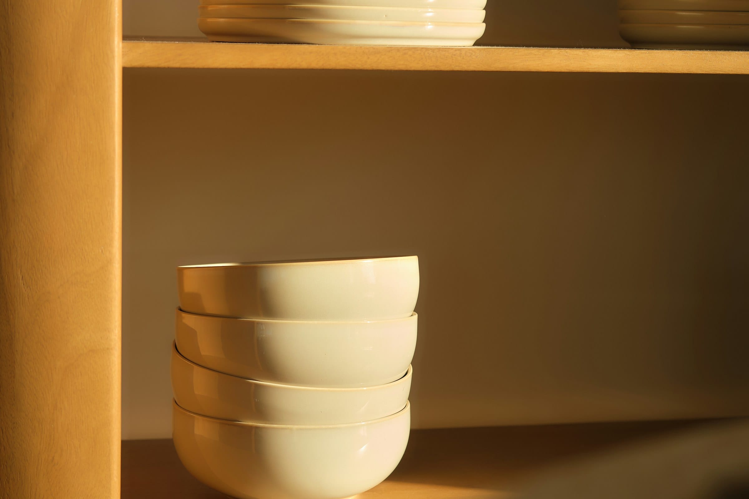 A column of cream glazed bowls catching low golden light on an oak shelf