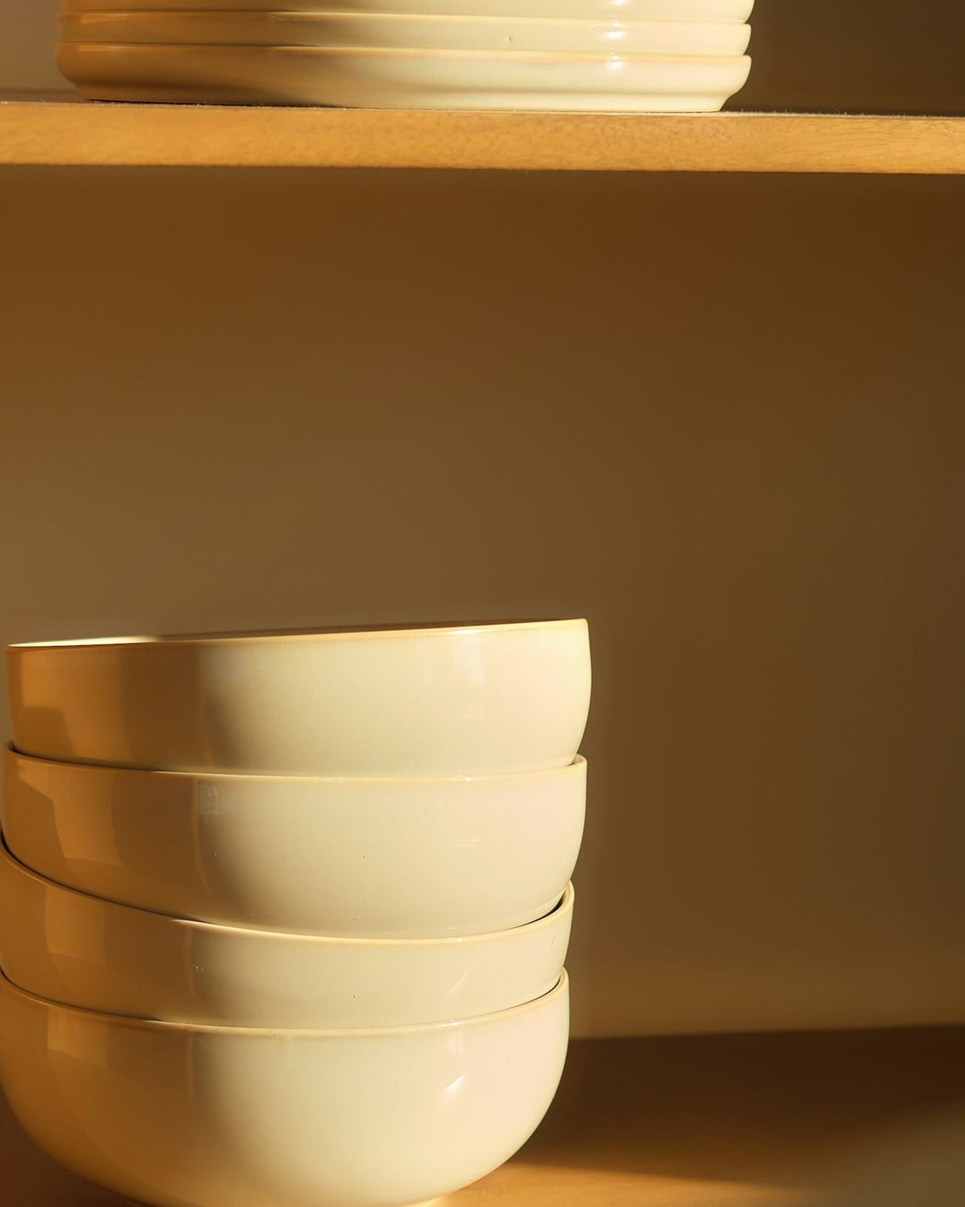 A column of cream glazed bowls catching low golden light on an oak shelf