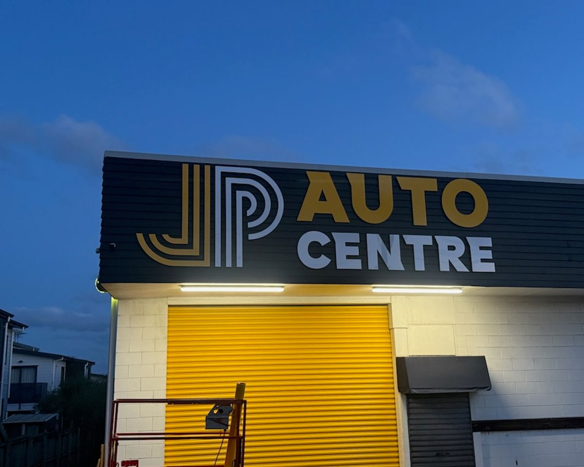 JP Auto Centre fascia sign — yellow JP letterform and AUTO CENTRE white text illuminated at twilight above a yellow workshop roller door