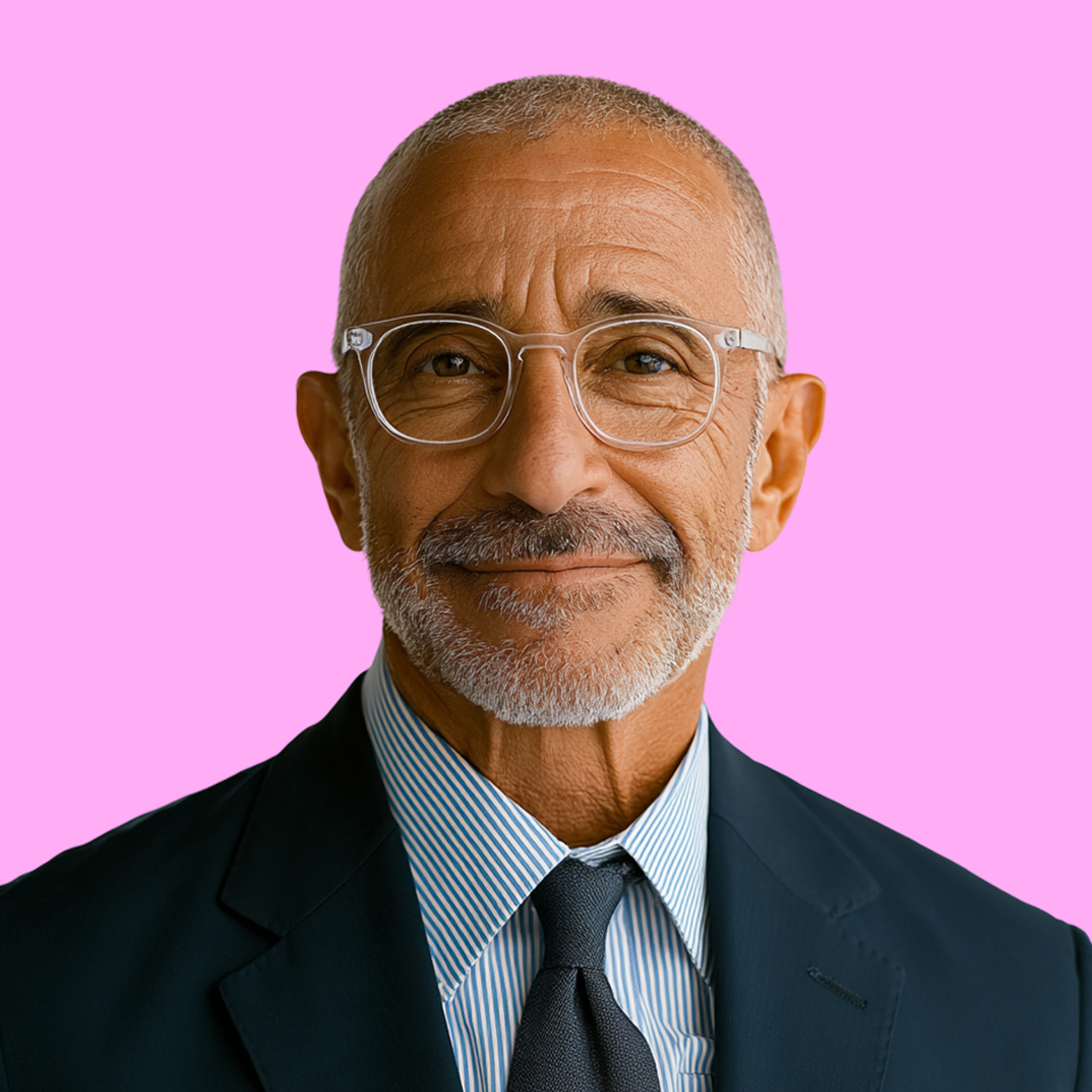 A smiling older man with gray hair, a beard, and glasses wears a suit and tie. He stands in front of a solid pink background.