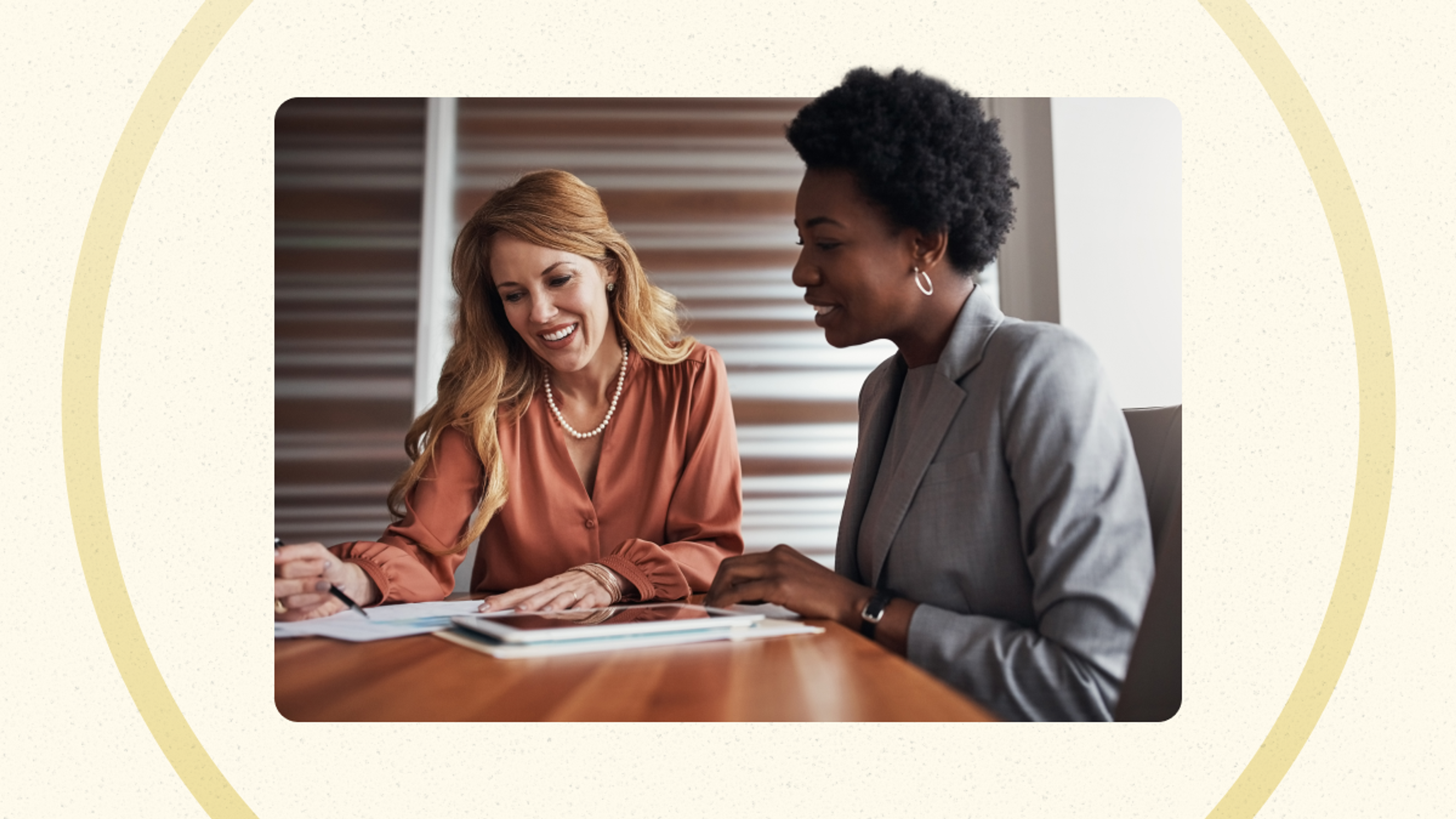 Two women sit at a table together, reviewing documents and smiling in a professional office setting.