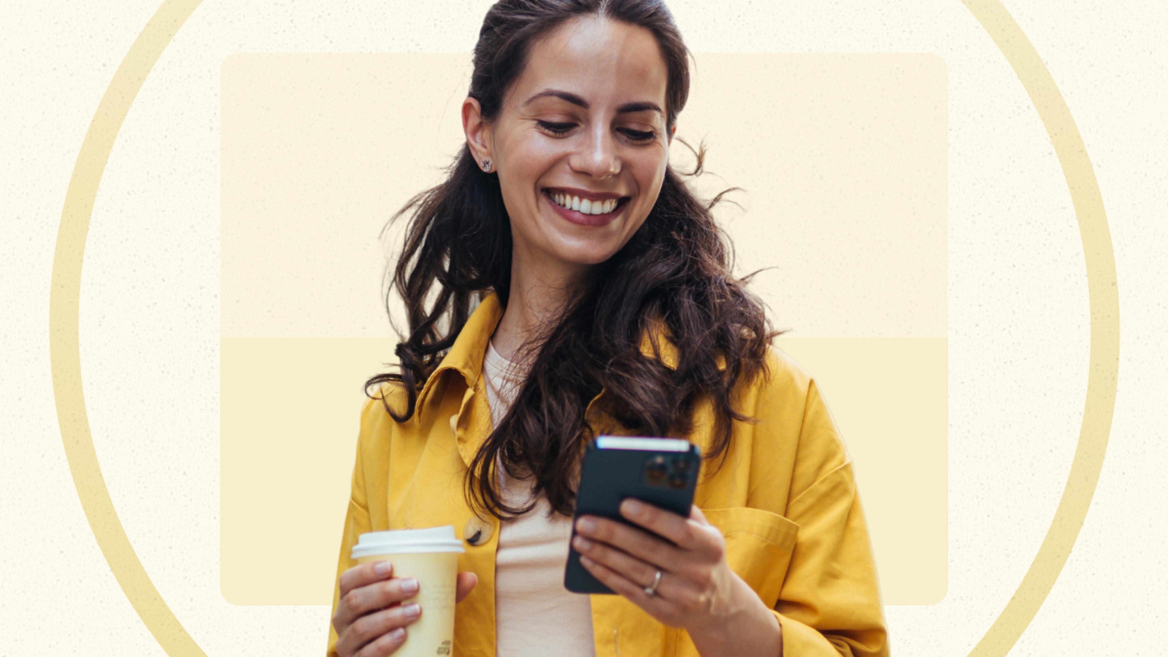 A woman in a yellow jacket holds a coffee cup and smiles while looking at her smartphone against a beige background.