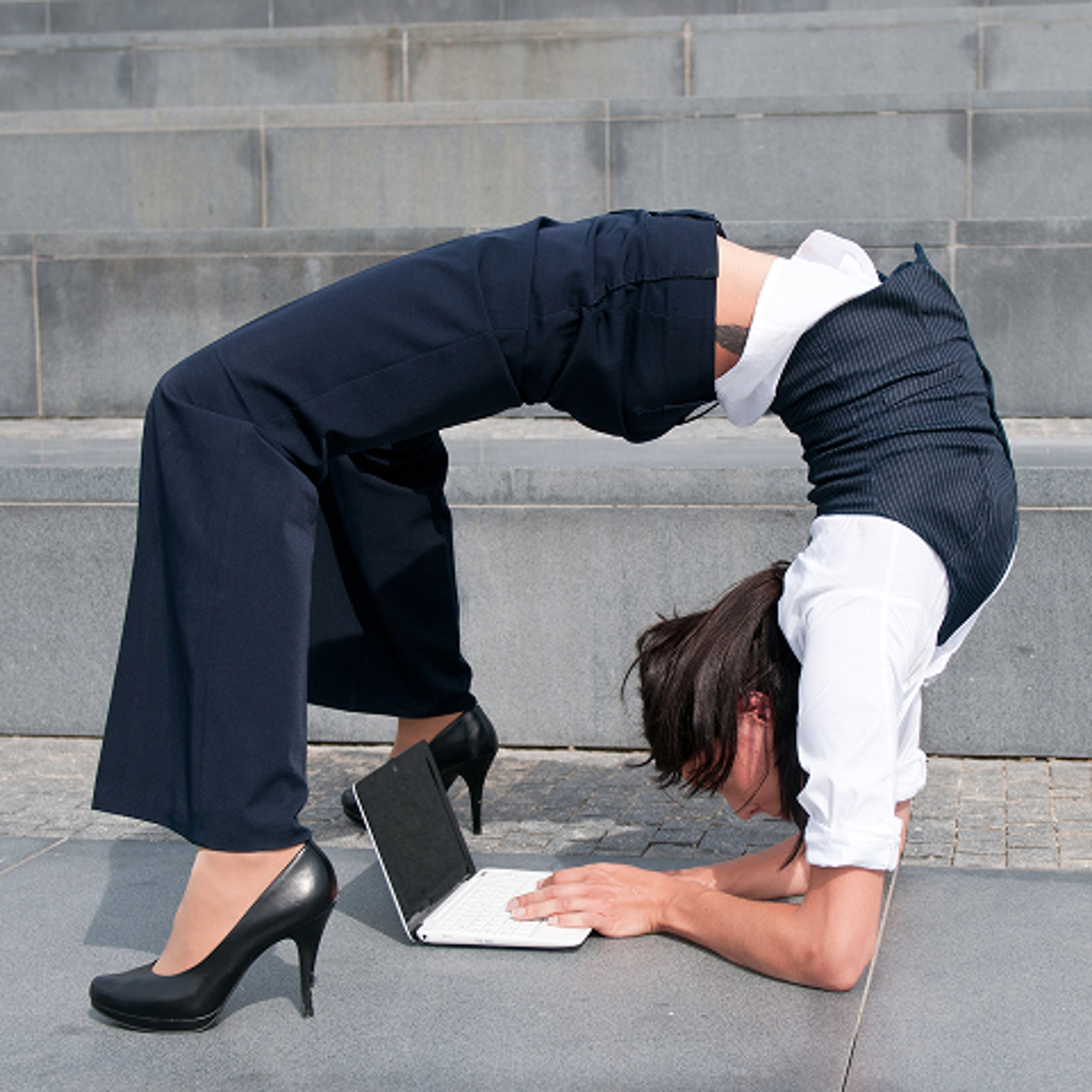 A woman in business attire performs a backbend on outdoor steps, balancing on her forearms and feet, while typing on a laptop placed on the ground in front of her.