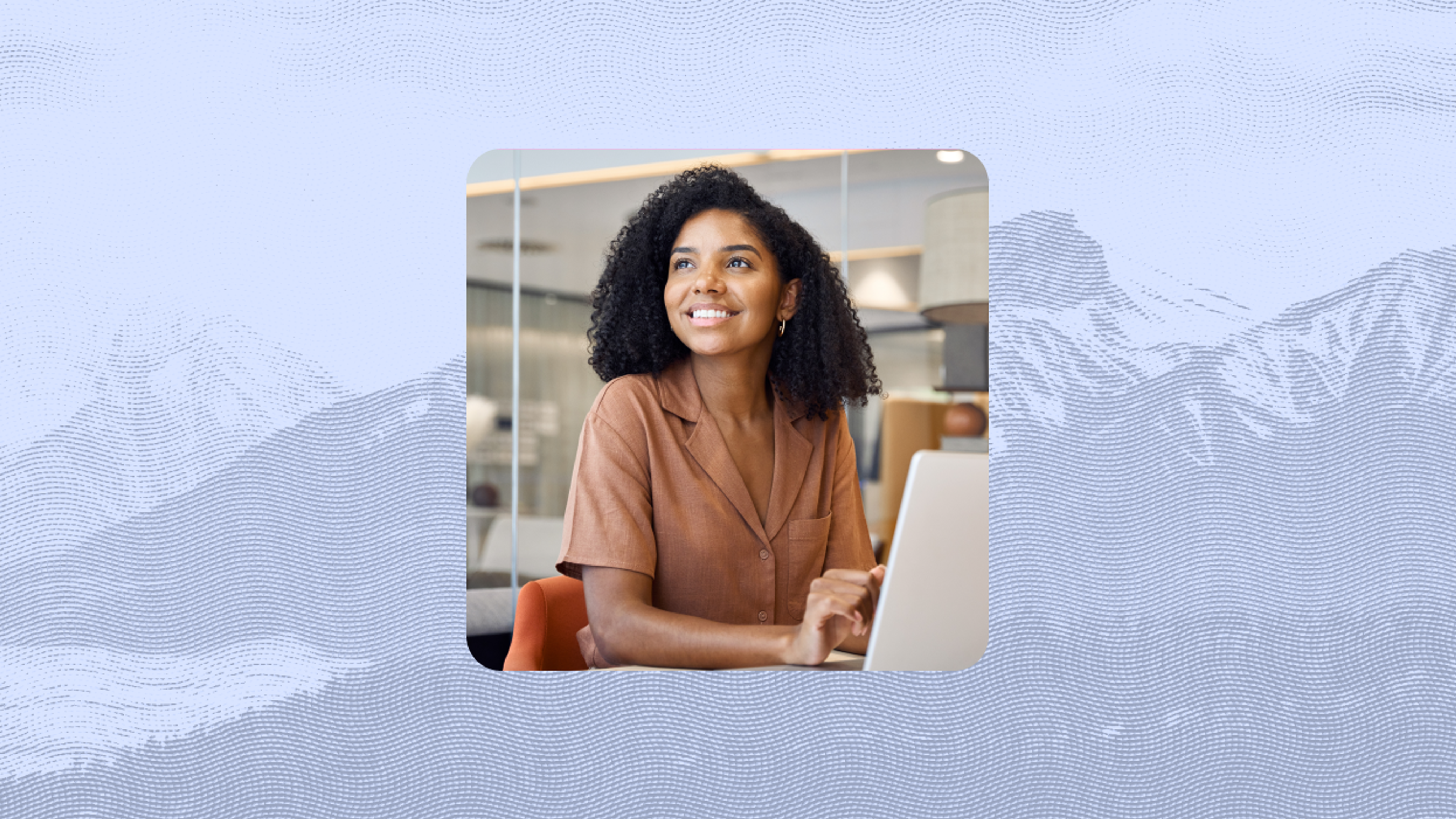 Woman with curly hair sits at a desk, smiling while working on a laptop in a modern indoor setting.