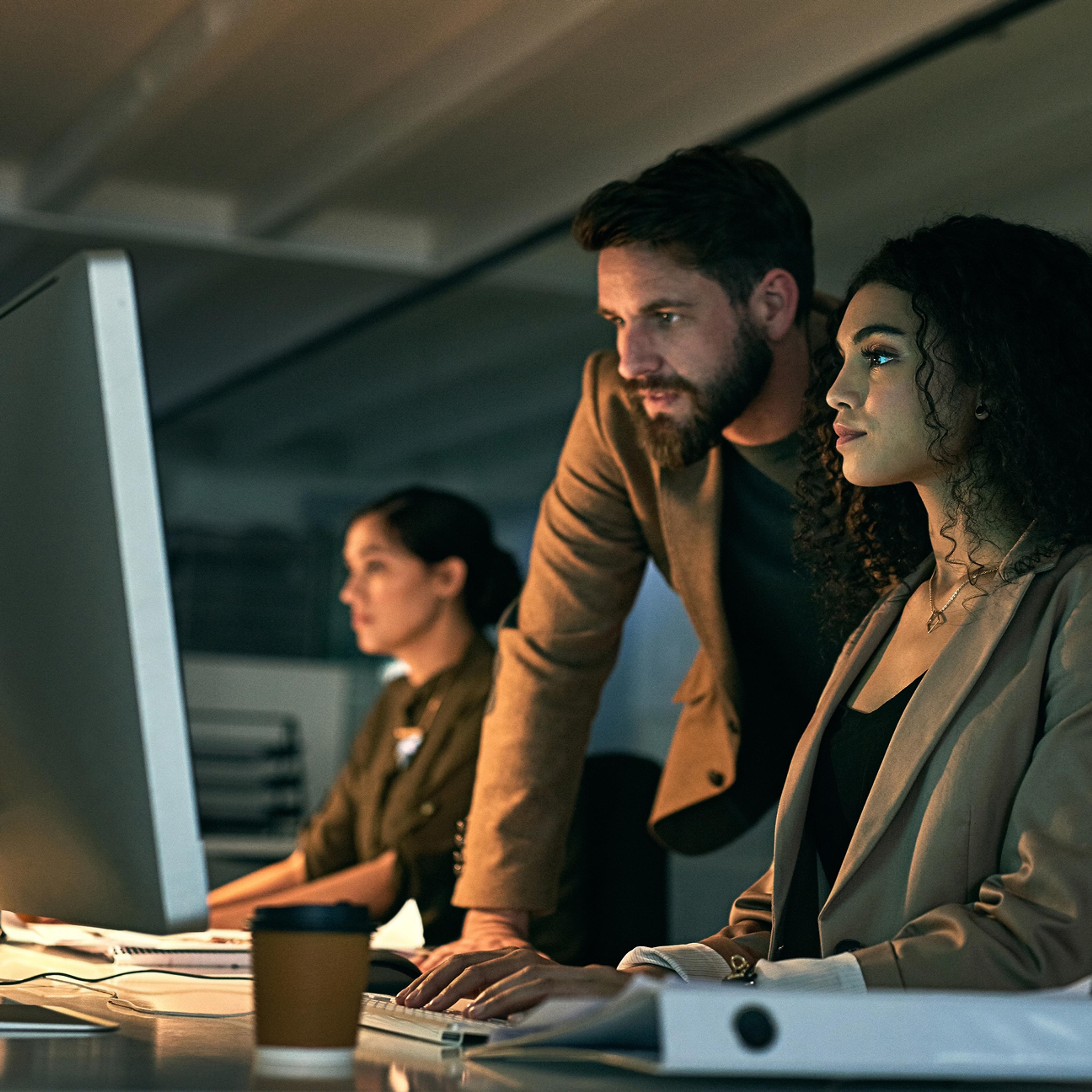 Two professionals, a man and a woman, work together at a computer in a modern office. Another woman works in the background. A coffee cup, files, and a keyboard are on the desk. The lighting is dim and focused.