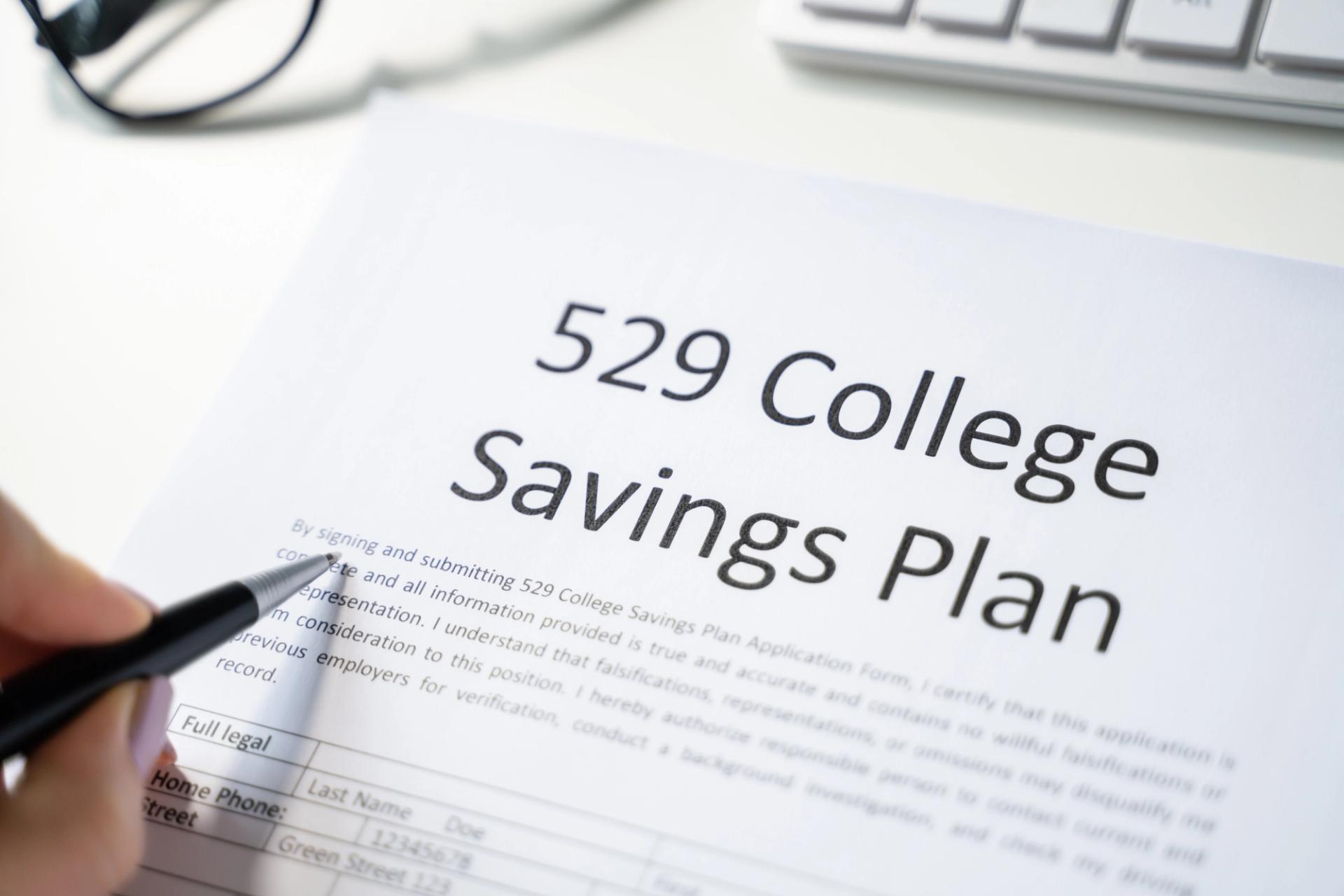 A person holding a pen reviews a document titled "529 College Savings Plan" next to a computer keyboard.