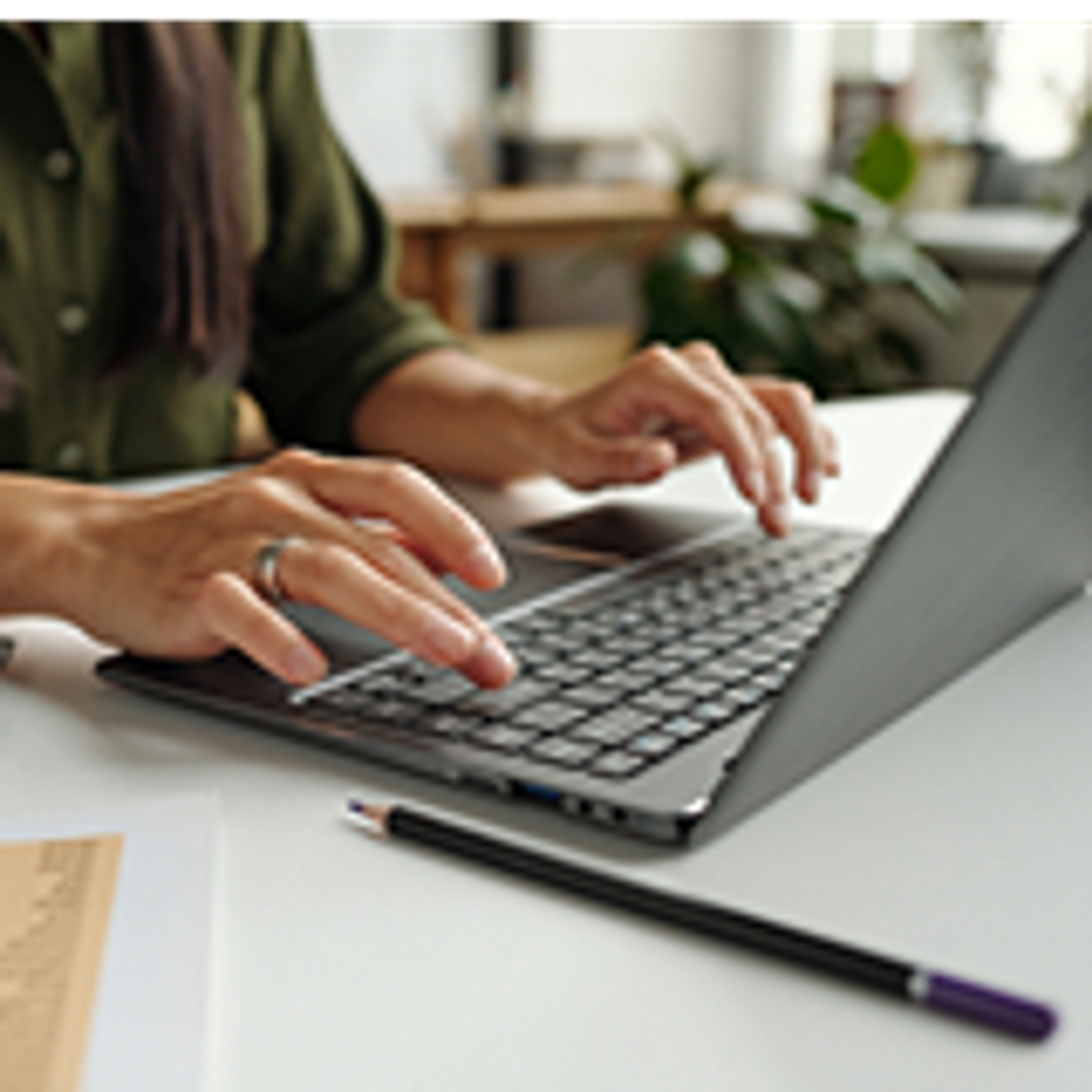 A person types on a laptop at a desk, with a pencil and documents nearby. The focus is on their hands and the keyboard; the background appears to be a bright, modern workspace.