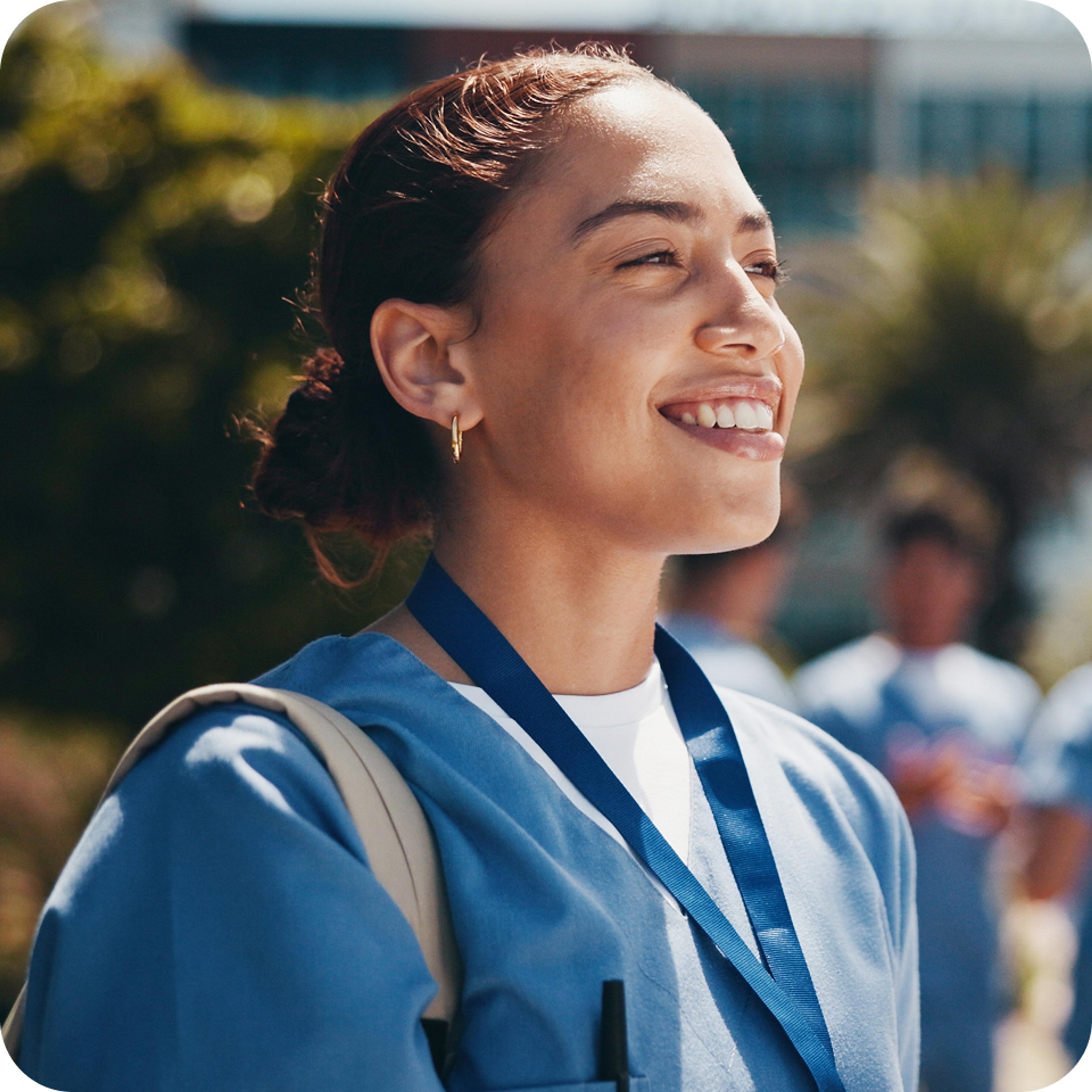 A smiling woman in a blue medical uniform, wearing a lanyard, looks upwards with a cheerful expression. The background shows a sunny outdoor setting with blurred figures, suggesting a positive and professional atmosphere.