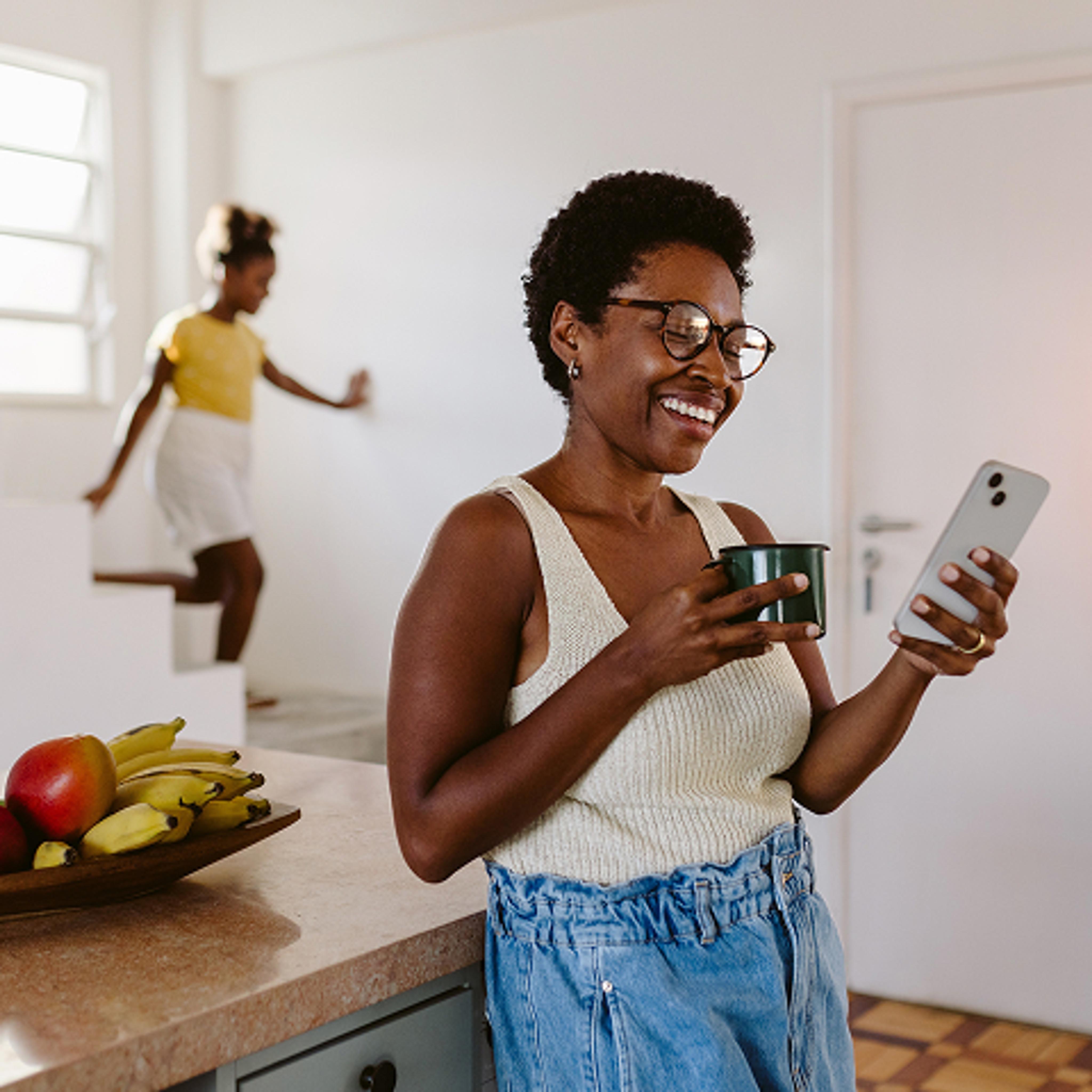 A woman in glasses smiles while looking at her smartphone and holding a mug in a bright kitchen. A bowl of fruit sits on the counter, and another person is coming down the stairs in the background.